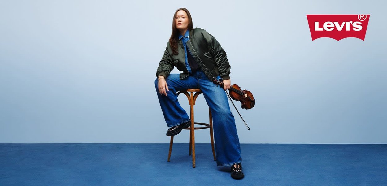 A person wears a green bomber jacket over a denim shirt and wide-leg blue jeans, holding a violin while sitting on a wooden stool.