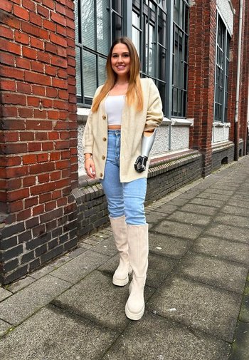 En kvinna står på en trottoar, iklädd en beige överdimensionerad cardigan, vit topp, ljusblå jeans och krämfärgade knähöga stövlar. Hon har en metallisk protesarm.