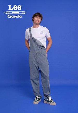 Striped denim overalls with one shoulder strap, worn over a white t-shirt. Pair with green and white sneakers. Background is solid blue.
