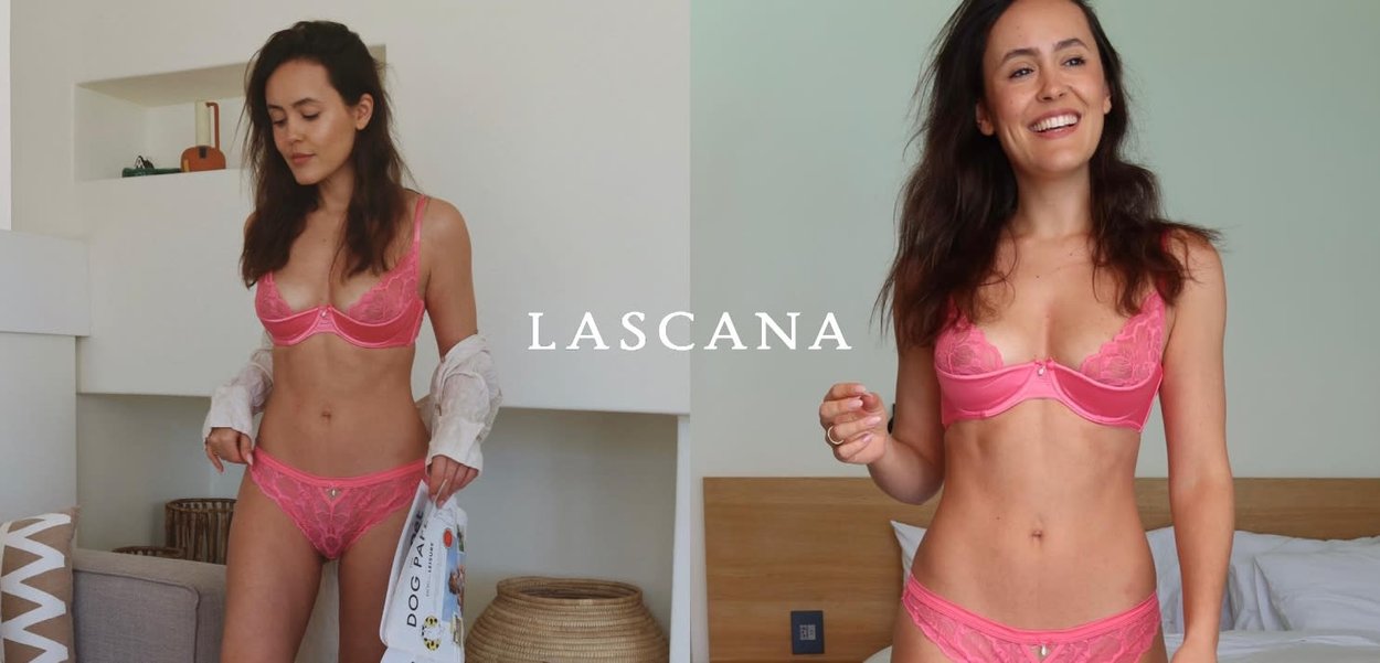 Frau in rosa Spitzenunterwäsche posiert und lächelt in einem hellen, modernen Schlafzimmer mit neutraler Dekoration.