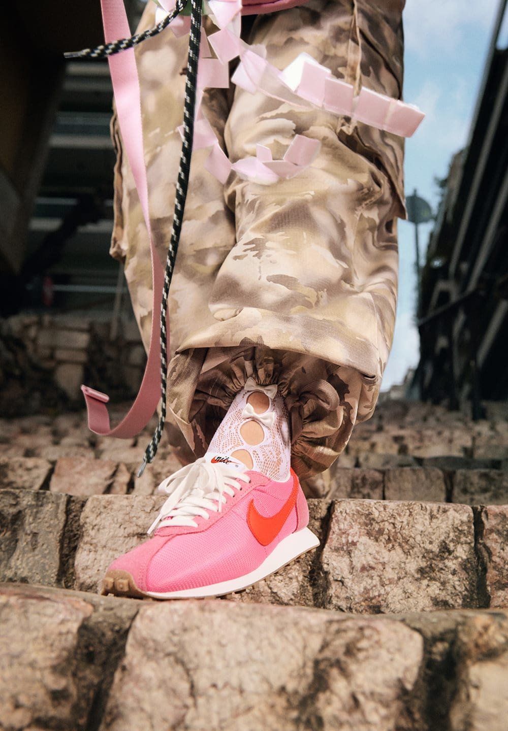 Nike sneakers with a pink mesh upper, orange swoosh, white sole, paired with camouflage pants and patterned white socks, on rocky steps.