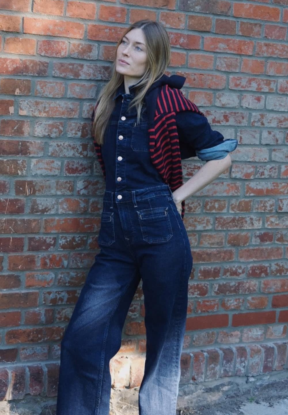 Denim overalls in dark blue with pockets and button front. Paired with a black and red striped hoodie draped over shoulders. Brick background.
