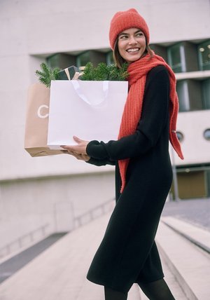 Robe noire côtelée à manches longues, bonnet rouge en tricot et écharpe. Tient des sacs de courses blancs et marron avec des feuillages. Cadre extérieur.