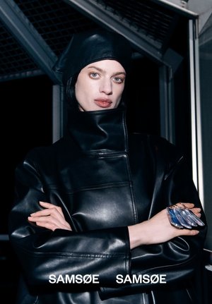 Persoon met een bleke huid en blauwe ogen, gekleed in een zwarte leren jas en bijpassende hoofddeksel, die verschillende transparante sleutelhangers of labels vasthoudt.