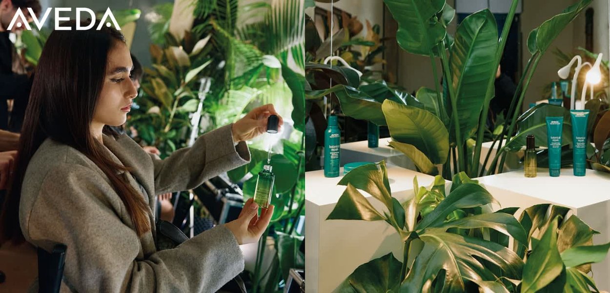 Groene glazen fles met druppelaar, grijze container en turquoise Aveda-producten op een witte display. Weelderige groene planten op de achtergrond.