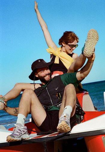 Due persone su una barca: una indossa un cappello a secchiello nero, un gilet nero e pantaloni corti; l'altra ha un fiocco giallo e occhiali da sole, mostrando un piede.