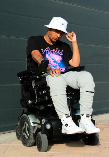 Elektrisk rullstol med svart ram, silverdetaljer och stora bakhjul. Användaren bär en svart grafisk t-shirt, grå sweatpants och vita sneakers.