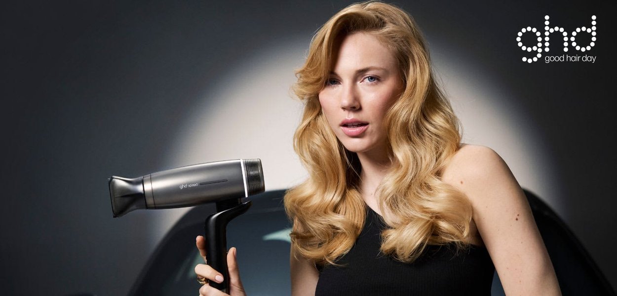 Blonde vrouw met volumineus golvend haar houdt een zwarte ghd-haardroger vast tegen een donkere gradient achtergrond met het ghd-logo.