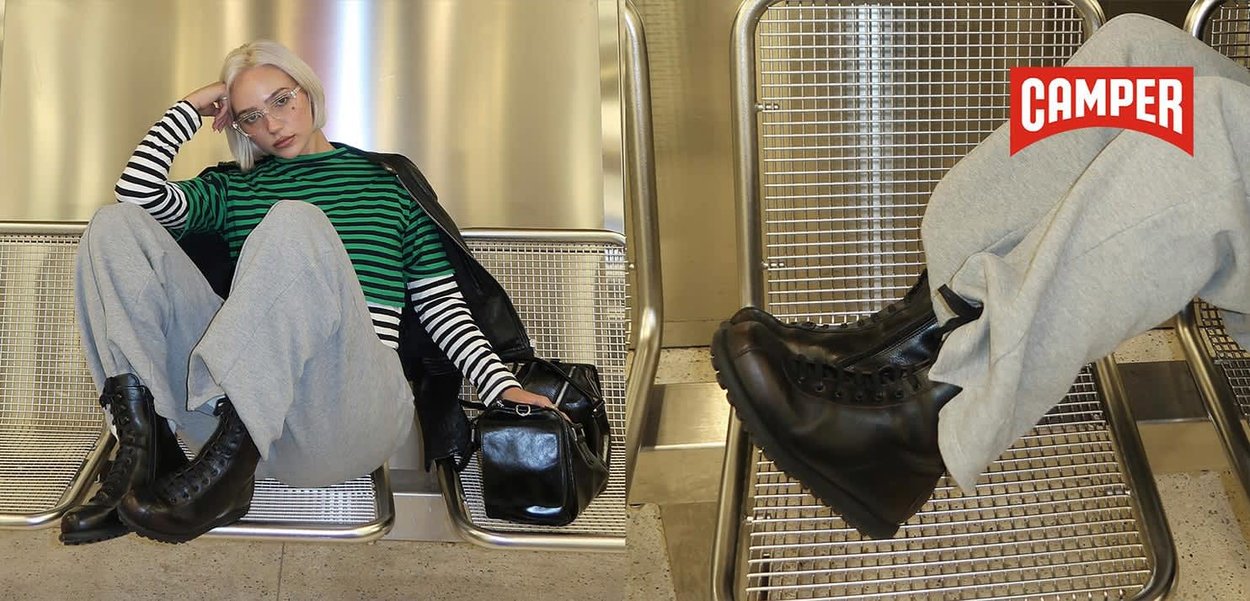Jeune personne avec des cheveux blonds courts, portant une chemise rayée, un pantalon gris et des bottes noires, assise sur un banc en métal, tenant un sac noir à l'intérieur.