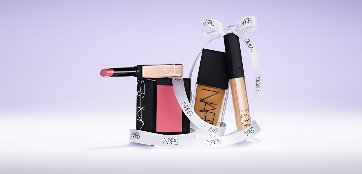 Set di cosmetici composto da un blush rosa compatto, un fondotinta liquido in un flacone di vetro, un rossetto cremoso e un nastro bianco con dettagli del logo.