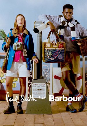 Zwei Models in lässiger Outdoor-Bekleidung stehen neben einem münzbetriebenen Fernrohr, halten Taschen und Requisiten auf einem Holzdeck mit einem Rettungsring im Hintergrund.