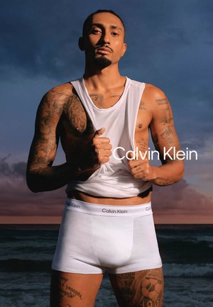 Uomo tatuato che solleva una canottiera bianca, indossa biancheria intima bianca Calvin Klein, in piedi su una spiaggia al tramonto con un cielo nuvoloso.