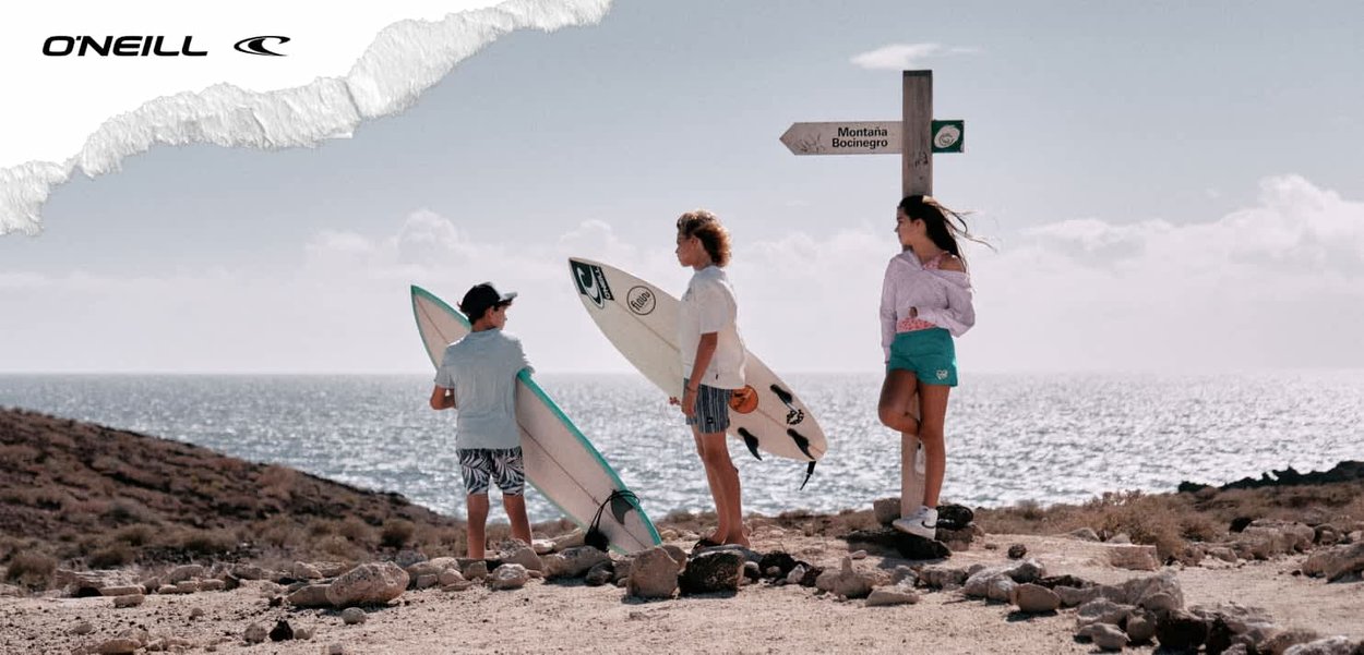 Zwei Buben halten Surfbretter und ein Mädchen steht neben einem Holzschild an einem felsigen Strand mit Ozean und bewölktem Himmel im Hintergrund.