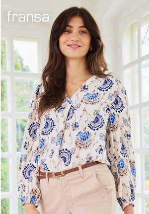 Frau trägt eine beige Blumenbluse mit blauen und braunen Mustern, die in eine hellbeige Hose mit braunem Gürtel gesteckt ist, steht drinnen am Fenster.