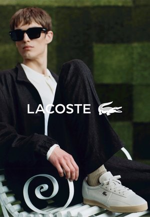 Białe sneakersy Lacoste ze skórą o teksturze, w połączeniu z ciemnymi materiałowymi spodniami. Model siedzi na białym kręconym krześle z zielonym tłem.