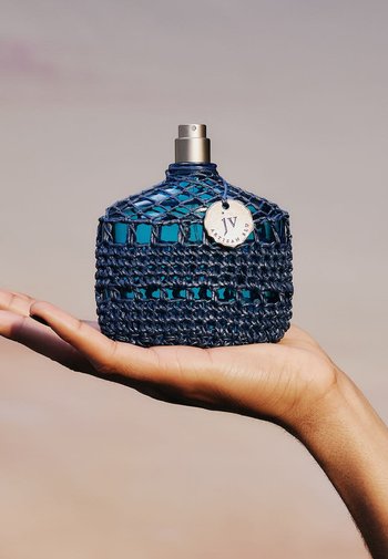 Bouteille de parfum en verre bleu avec texture tissée marine, vaporisateur en argent, et une étiquette marquée "Artisan Blu." Tenue dans une main ouverte.