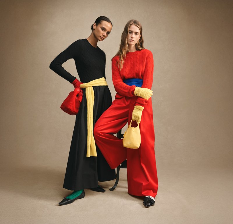 Zwart gebreide jurk met lange mouwen, gele ceintuur in de taille, rode kabelgebreide trui, wijde rode broek, gele clutch, groene sokken, zwarte schoenen.