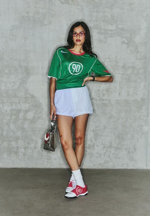 Groene trui met witte accenten en een rond logo, gecombineerd met lichtblauwe shorts, rode sneakers en een doorzichtige tas met een embleem.