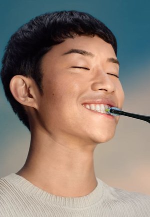 Giovane con capelli neri corti che sorride con gli occhi chiusi mentre si spazzola i denti superiori con uno spazzolino elettrico nero.