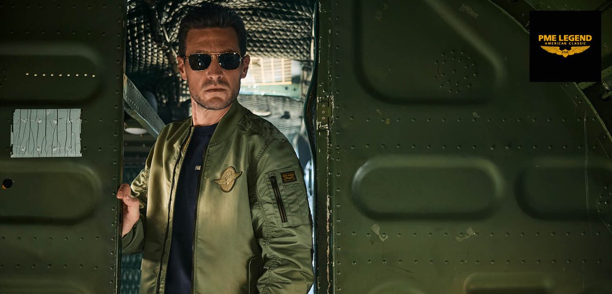 Hombre con gafas de sol y una chaqueta tipo bomber de color verde oliva está en la puerta de una aeronave de estilo militar.