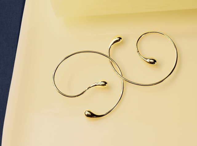 Δύο κομψά, λεπτά χρυσά βραχιόλια ανοικτού τύπου σε σχήμα cuff, τοποθετημένα σε μια λεία μπεζ επιφάνεια με μια σκοτεινή μπλε γωνία.