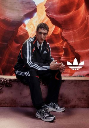 Jonge man in een zwart Adidas trainingspak en zilveren sneakers zittend in een rood rotsachtige canyon met het Adidas-logo zichtbaar naast hem.