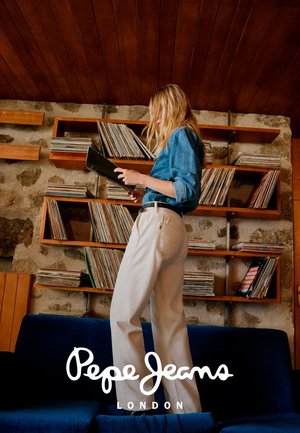 Helles jeanshemd in Blau, beige weit geschnittene Hosen, stehend vor Holzregalen mit Vinyl-Schallplatten. Dunkelblaues Sofa im Vordergrund.