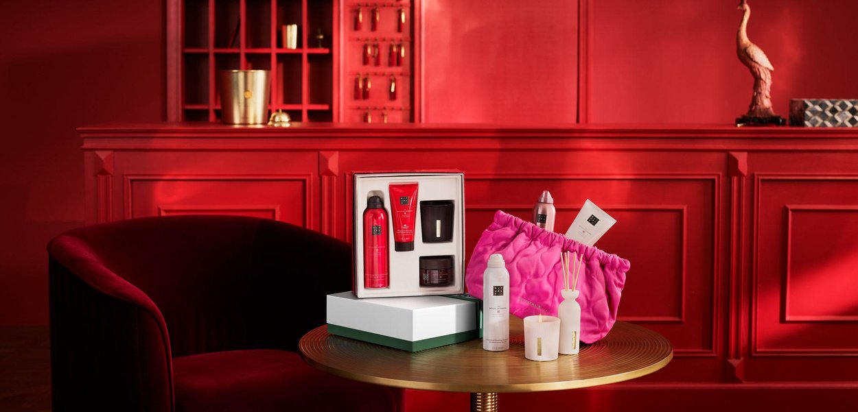 Set includes a white and green box, a pink padded bag, red and black containers, a candle, and a fragrance diffuser on a round gold-striped table.