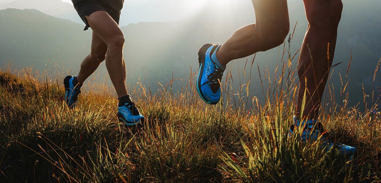 Deux coureurs en chaussures de sport bleues courent à travers de l'herbe haute sur une colline, mettant en valeur le matériau de la chaussure et sa semelle de traction.