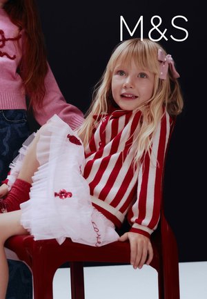 Rood-wit gestreepte trui, laagjes tule rok met bloemmotieven, gezeten op een rode stoel, tegen een donkere achtergrond.