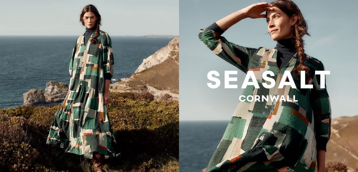 Kvinde iført en flerfarvet patchwork-kjole og sort turtleneck, stående på en græsbelagt klippe med udsigt over havet under en klar himmel.