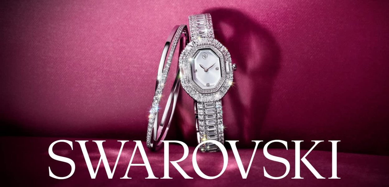 Zilveren horloge met een octagonaal wijzerplaat, kristallen versieringen en een metalen band. Vergezeld van een bijpassende zilveren armband. Roze achtergrond.