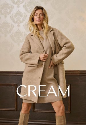 Manteau en mélange de laine beige avec de grands boutons, tissu texturé et coupe oversize, superposé à une robe assortie, accompagné de bottes till genou.
