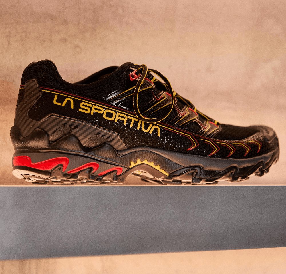 Black trail running shoe with yellow and red accents, textured mesh upper, rugged sole, and a distinct tread pattern for traction.