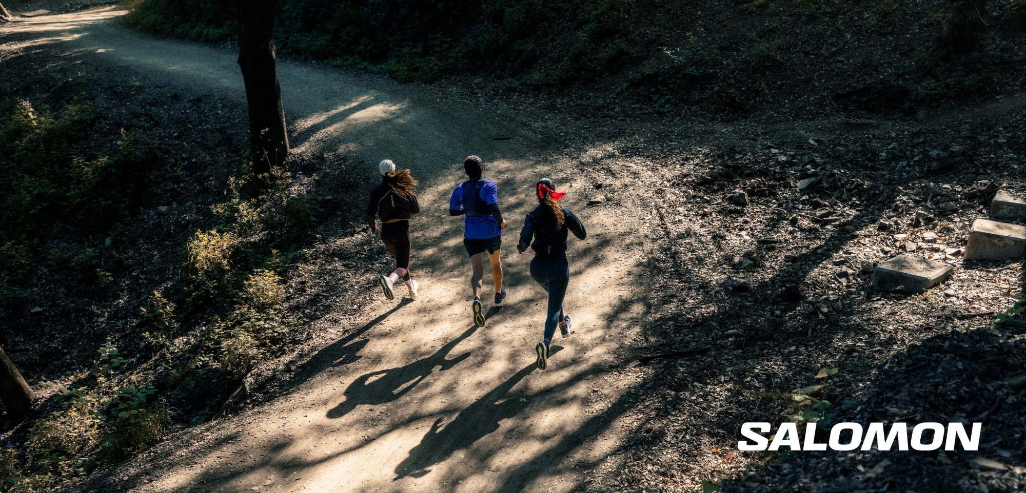 Trois coureurs sur un chemin en terre, portant des vêtements de sport variés en couleurs sombres et vives. La scène présente des ombres et un arrière-plan boisé.