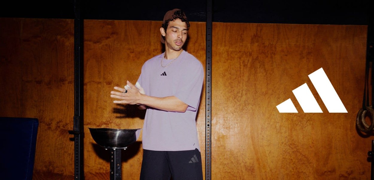 T-shirt de sport violet avec des accents noirs et un logo, associé à un short noir. Le sujet tient de la craie, se tenant dans une salle de sport contre un mur en bois.