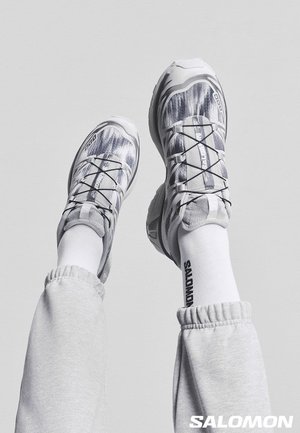 Gambe sollevate che indossano scarpe da corsa Salomon bianche e grigie con motivo, calze bianche con il testo "Salomon" e pantaloni della tuta grigio chiaro su uno sfondo grigio.