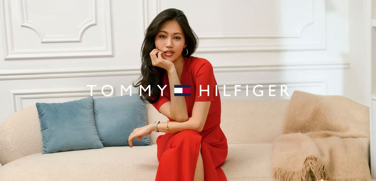 Mujer con vestido rojo sentada en un sofá beige con cojines azules, posando pensativamente en una habitación minimalista con paneles blancos.