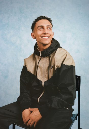 Zwart en beige windbreaker met een rits aan de voorkant en capuchon, voorzien van elastische manchetten. Draagt een bijpassende zwarte broek. Achtergrond is lichtblauw.