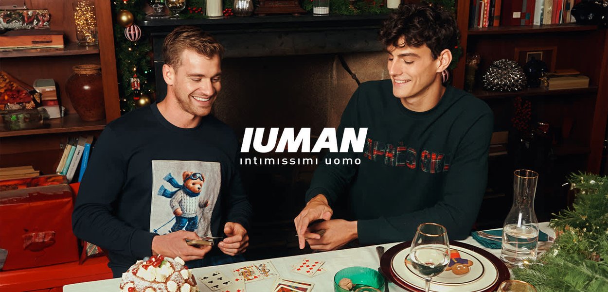 Deux hommes assis à une table portant des pulls, l'un avec un motif d'ours, l'autre en vert foncé. Ils jouent aux cartes dans un décor et une ambiance festive.