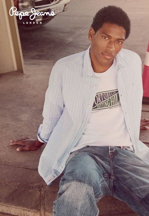 Man zittend op de betonnen vloer, gekleed in een lichtblauw gestreept overhemd over een wit grafisch T-shirt en een blauwe spijkerbroek, met de handen op de grond achterover leunend.