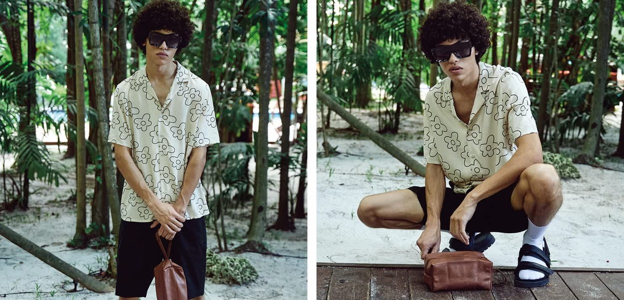 Mann mit lockigem Haar, der eine Sonnenbrille, ein geblümtes Hemd, schwarze Shorts und Sandalen trägt, posiert stehend und hockend und hält eine braune Tasche in einem Wald.