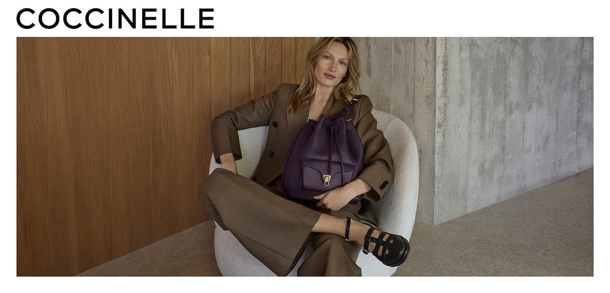 Abito marrone con spalline, borsa in pelle viola con chiusura a coulisse e tasca frontale, sedia rotonda bianca, sfondo in legno e cemento.