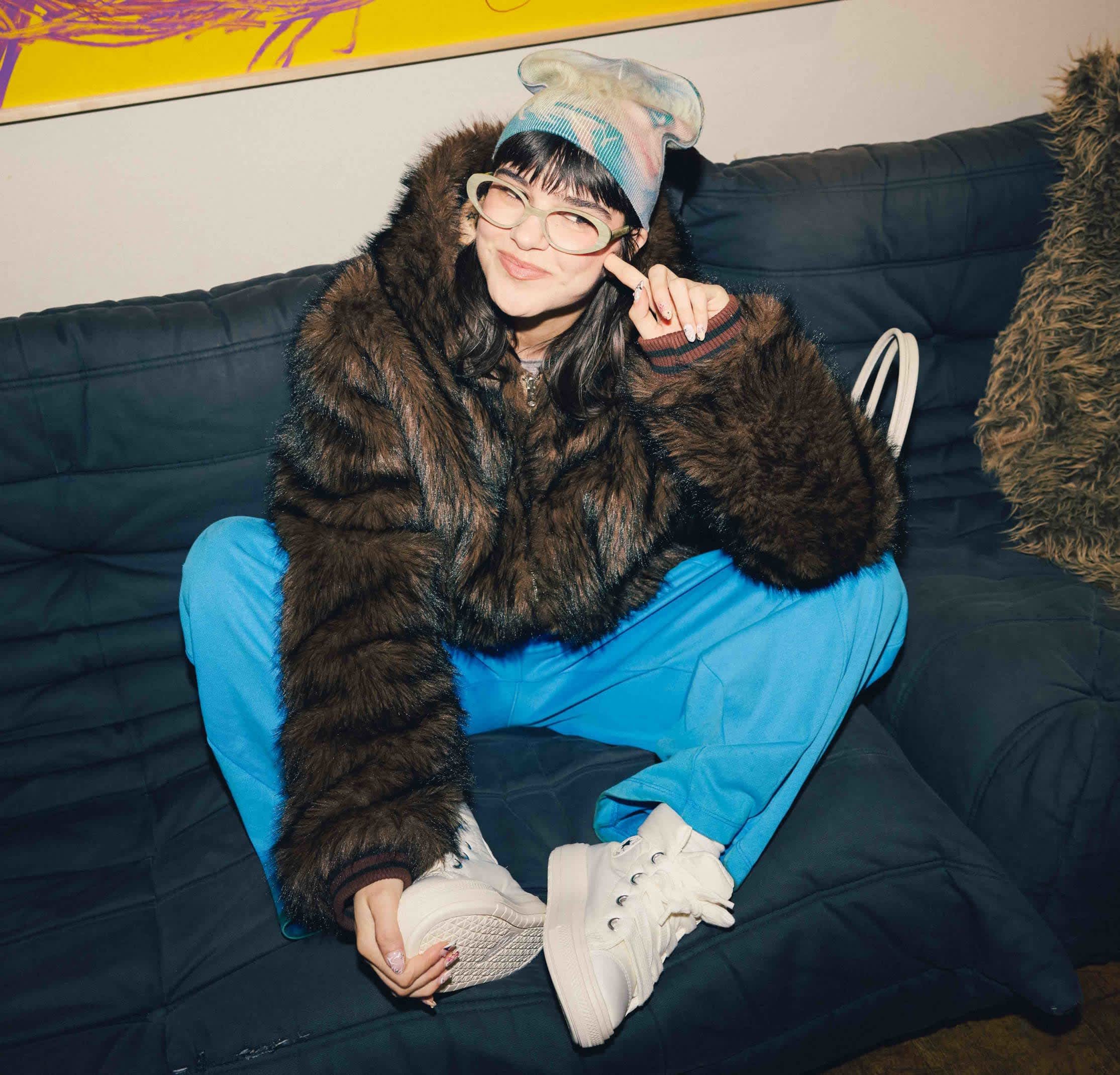 Sudadera con capucha de piel sintética marrón, pantalones azules y zapatillas blancas con plataforma. Gafas y pañuelo colorido con estampado. Sentada en un sofá oscuro.