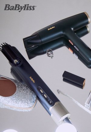 Svart og grønn hårføner og stylingverktøy med knapper og børster, sammen med en sølvglitrende sko og et lite svart objekt, på en reflekterende overflate.