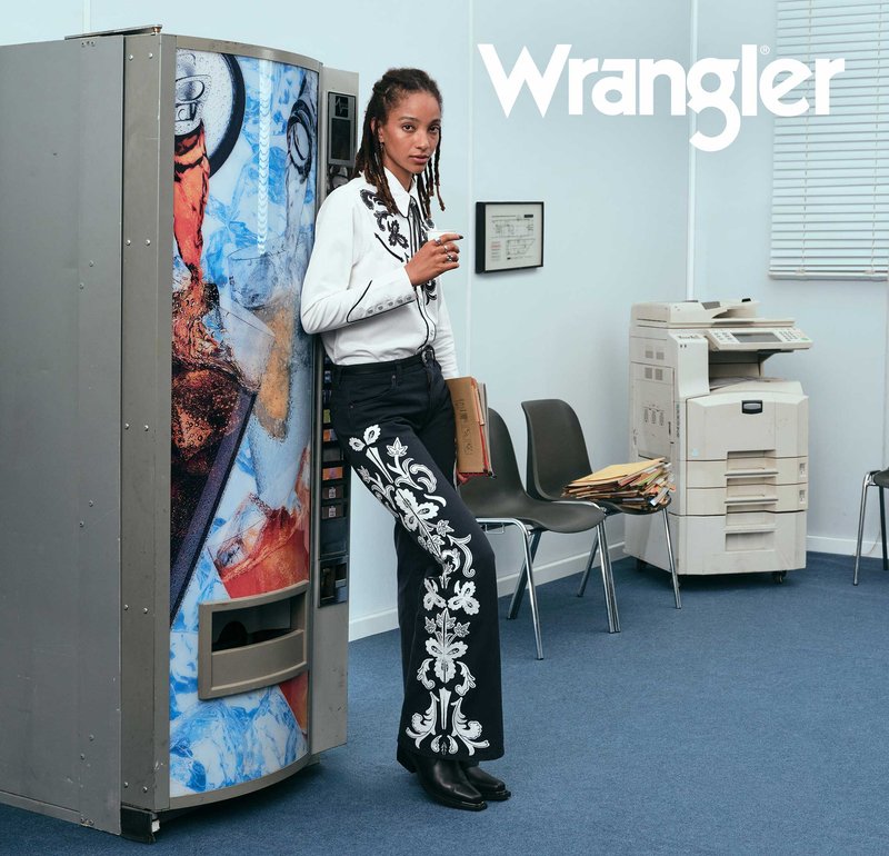 Biała koszula z guzikiem i czarnym haftem, czarne spodnie z białym kwiatowym wzorem, czarne botki, automat do napojów oraz drukarka w biurowym otoczeniu.