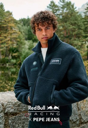 Mann mit lockigem Haar, der eine marineblaue Fleecejacke mit den Logos von Red Bull Racing und Pepe Jeans trägt, draußen neben Bäumen und einer Steinmauer.