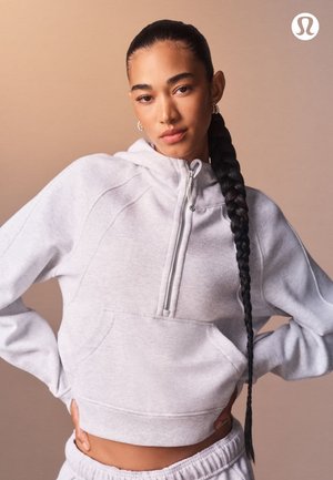 Hellgrauer, kurz geschnittener Hoodie aus weichem Stoff, mit halbhohem Reißverschluss vorne, Seitentaschen und gestickten Designdetails. Durchgehend glatte Textur.