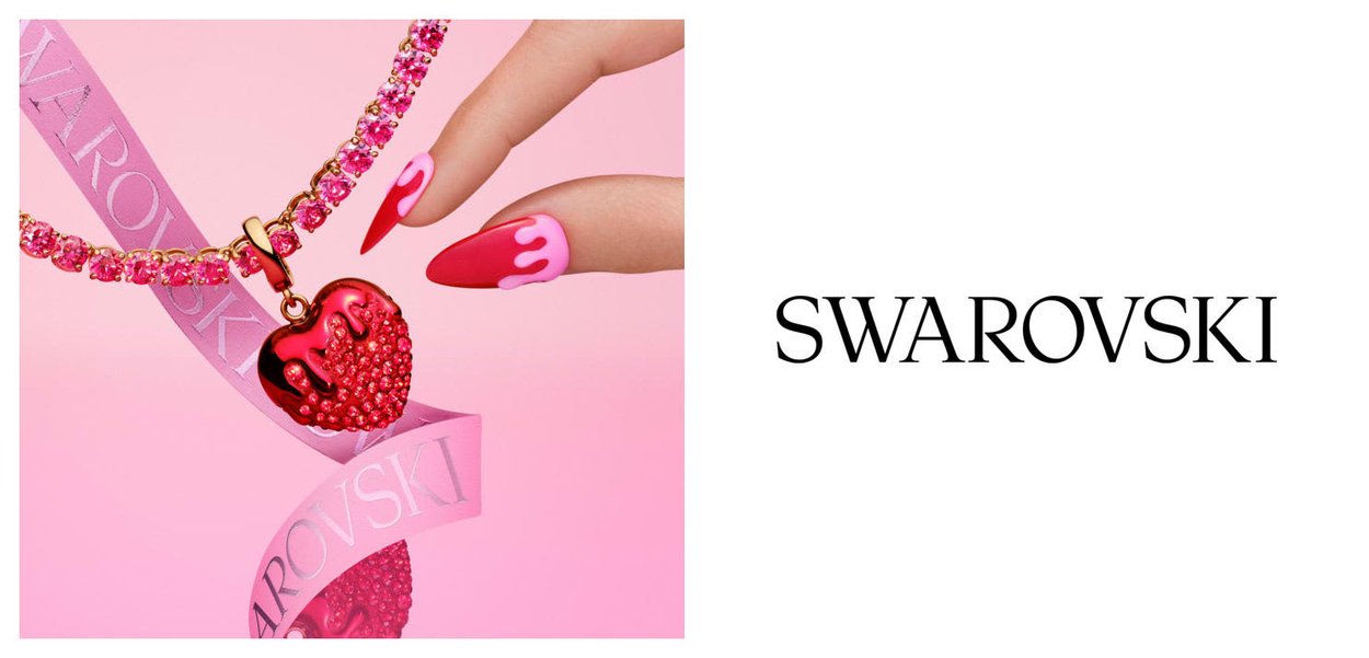 Main tenant un bracelet en cristal rose avec un pendentif en forme de cœur rouge et un ruban Swarovski rose sur un fond rose.