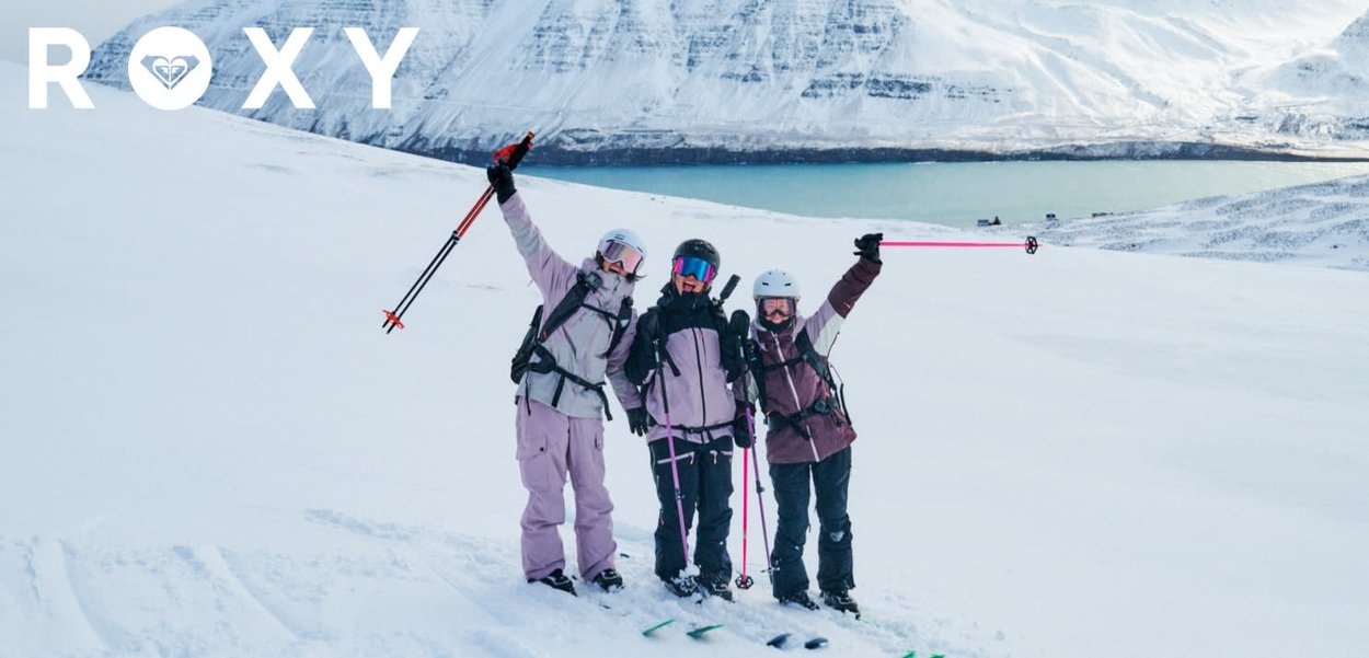 Trois skieurs dans la neige, portant des vestes violettes et noires, avec des bâtons de ski. Paysage enneigé et un plan d'eau en arrière-plan.
